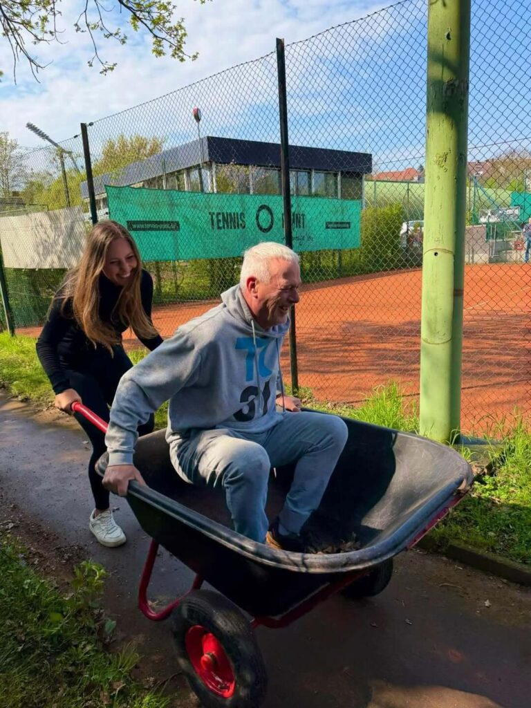 Ein Vereinsmitglied des TC31 Kassel wird von einem anderen Helfer lachend in einer Schubkarre über die Anlage geschoben.