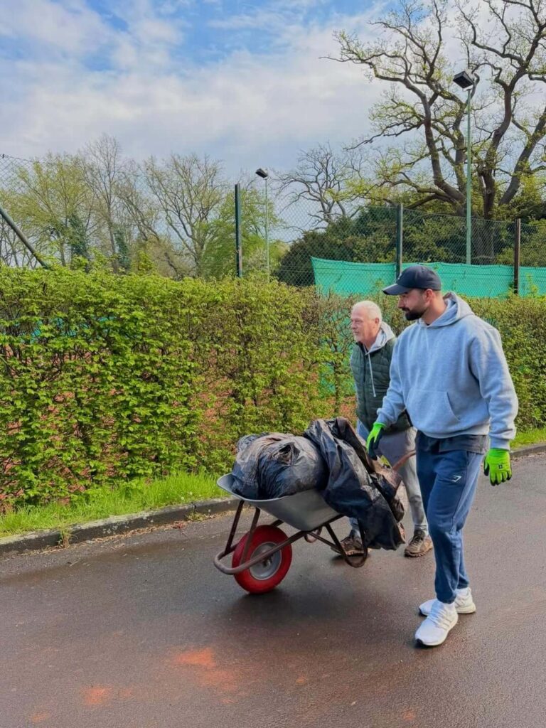 Zwei Männer transportieren Grünschnitt und Gartenabfälle in einer Schubkarre über die Tennisanlage.