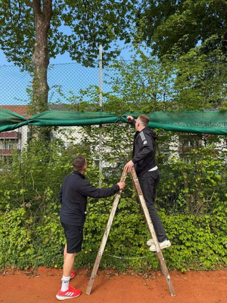 Zwei Männer auf einer Leiter befestigen die grünen Sichtschutznetze am Zaun des Tennisplatzes.